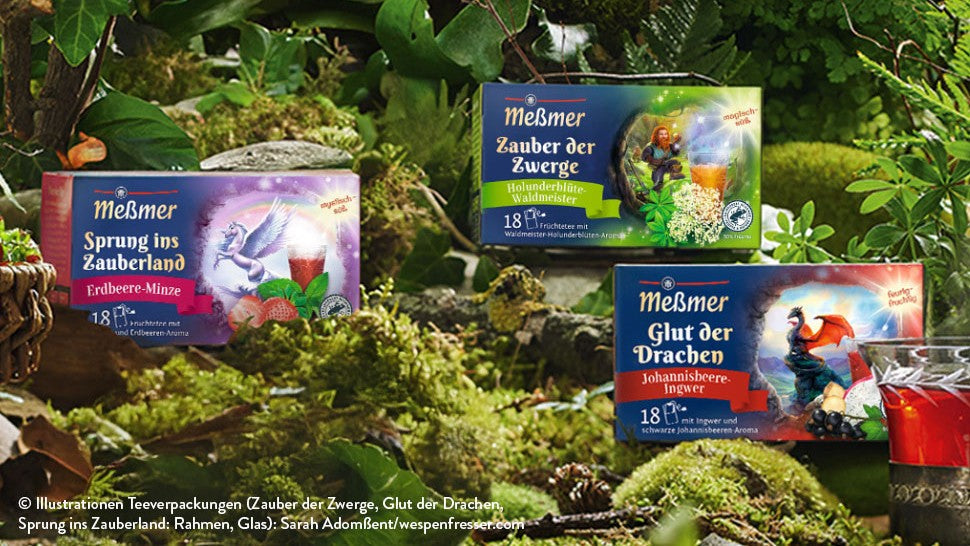 Zwei Schachteln Meßmer-Tee mit fantasievollen Motiven stehen in einer üppigen, grünen Waldlandschaft neben einer Glastasse mit leuchtend rotem Tee. Moos und Blätter umgeben die Szene.