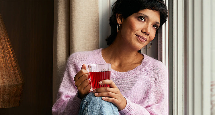 Eine Frau in einem hellrosa Pullover sitzt am Fenster und hält einen mit roter Flüssigkeit gefüllten Glasbecher in der Hand. Sie blickt nachdenklich hinaus und stützt ihren Arm auf ihr Knie, wobei ihr Gesicht von sanftem Licht erhellt wird.