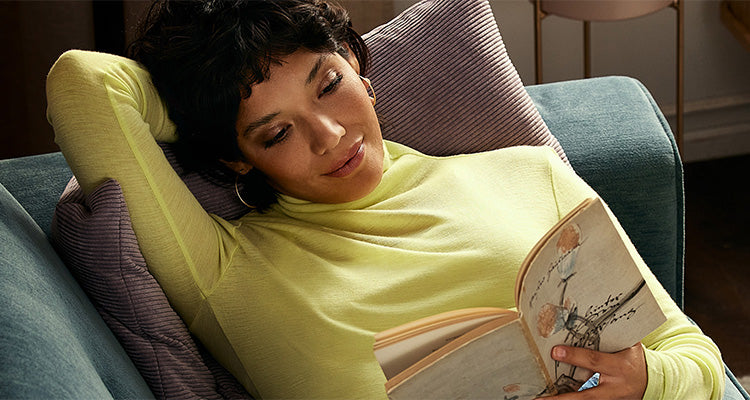 Eine Frau in einem gelben, langärmeligen Oberteil liegt entspannt auf einer Couch, lehnt sich mit einem Arm hinter dem Kopf an ein Kissen und liest mit einem sanften Lächeln ein Buch mit illustriertem Einband.