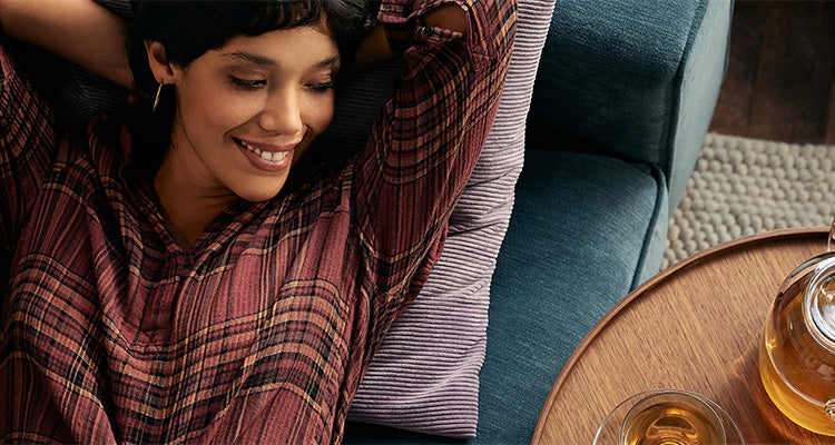 Eine Frau im karierten Hemd lächelt und entspannt sich auf einer blauen Couch mit den Armen hinter dem Kopf, neben einem runden Holztisch mit einer Teekanne und einem Glas Tee.