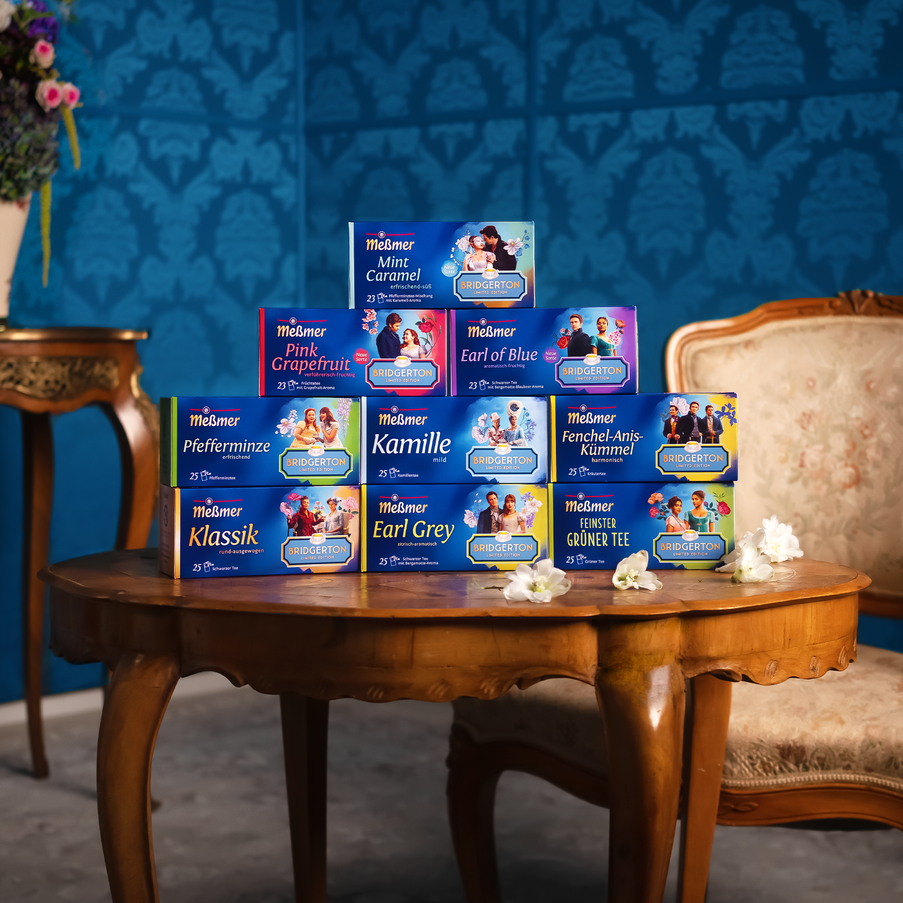 Mehrere Teepackungen, darunter Mint Caramel, arrangiert auf einem Holztisch vor blauem Hintergrund und antiken Möbeln in elegantem Innenraum.