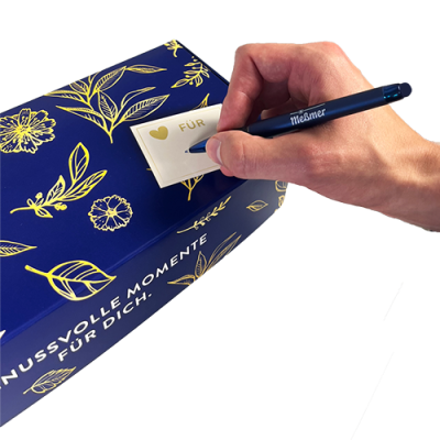 Eine Hand schreibt auf einen "FÜR"-Zettel mit goldenem Herz, der an der mit Tee gefüllten, floral gemusterten Geschenkbox Frühling von Meßmer DE befestigt ist und auf dem "GENUSS VOLLE MOMENTE FÜR DICH" steht.