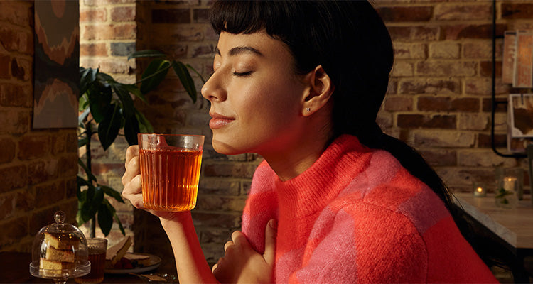 Eine Frau in einem rosafarbenen Pullover sitzt mit geschlossenen Augen und einem Lächeln im Raum, während sie eine Glastasse mit Tee an ihr Gesicht hält. Im Hintergrund sind ein kleiner Kuchen unter einer Glaskuppel und eine Topfpflanze zu sehen.