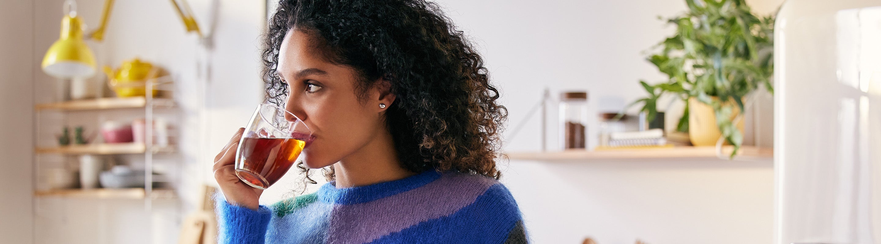Eine Frau mit lockigem Haar und einem bunten Pullover trinkt Tee aus einer Glastasse in einer hellen, modernen Küche mit Pflanzen und Regalen im Hintergrund.