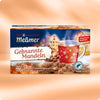 Eine Schachtel Meßmer DE Gebrannte Mandel Tee wird mit einer Glastasse mit rotem Tee, Schneeflockenmotiven, gerösteten Mandeln und einem festlichen Winterhintergrund gezeigt. Die Schachtel enthält 20 Rooibos-Teebeutel mit Mandelaroma.