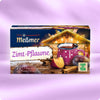 Wintertee Bestseller Set mit Meßmer Zimt-Pflaume, Teetasse, frischen Pflaumenstücken und Zimtstangen vor gemütlich beleuchtetem Haus auf lila Hintergrund