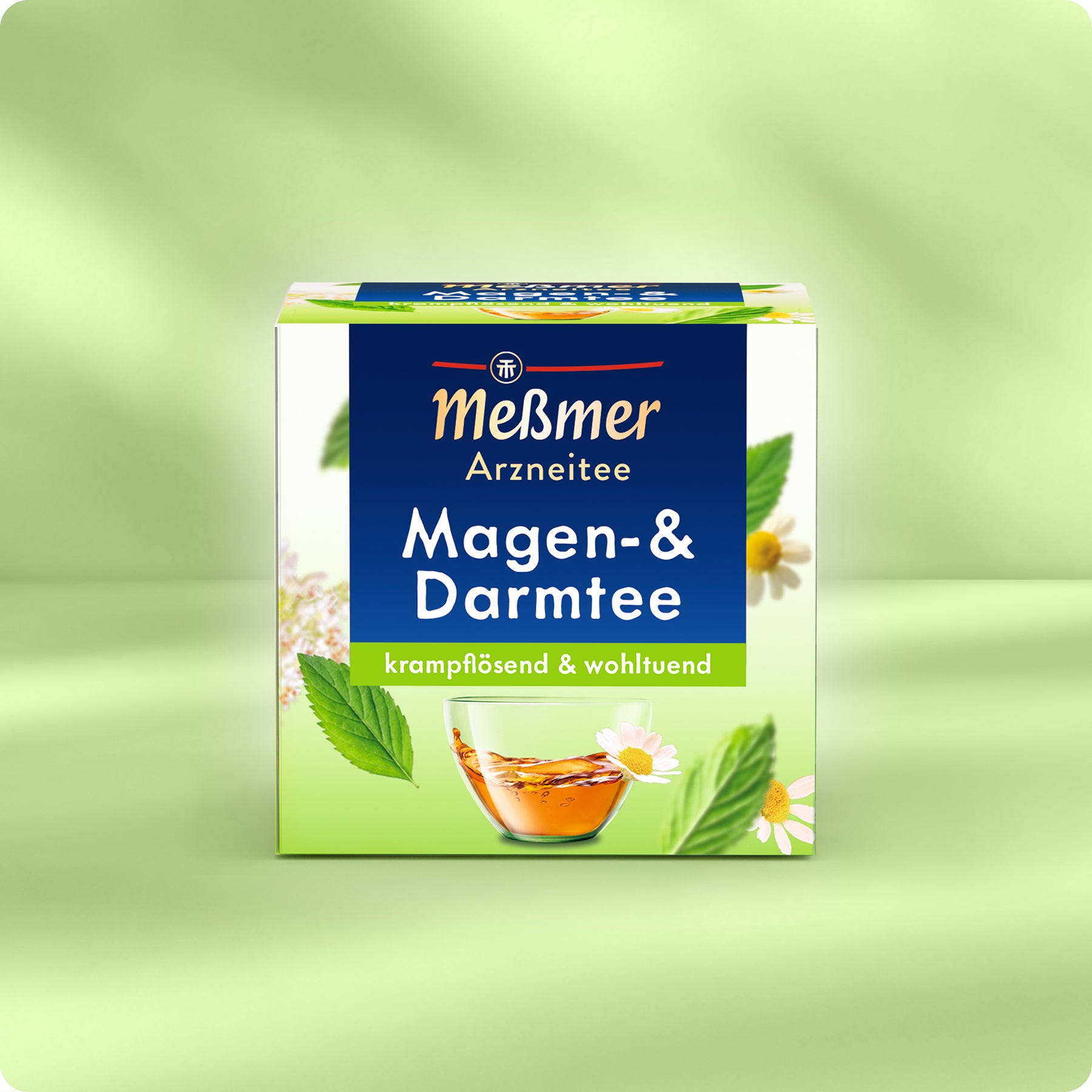 Eine Schachtel Meßmer DE Magen- und Darmtee, ein pflanzliches Heilmittel für Verdauungsprobleme, steht auf einer hellgrünen Fläche mit Kamillenblüten, grünen Blättern und einer Teetasse auf der blau, grün und weiß gestalteten Verpackung.