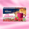 Verpackung von Heiße Himbeere mit dampfender Tasse Früchtetee, Himbeeren und Care-for-Tea-Siegel vor festlich beleuchtetem Hintergrund für nachhaltige Zutaten.