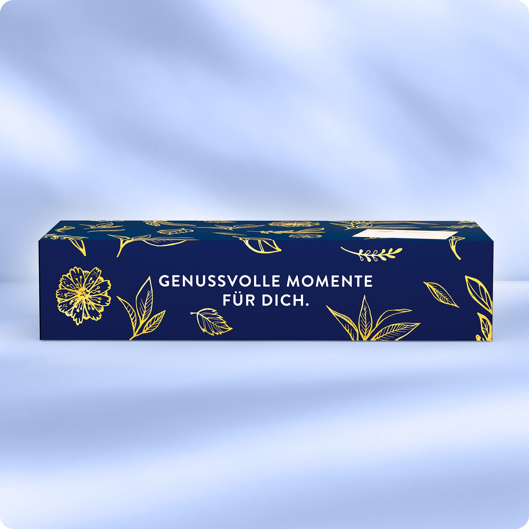 Eine rechteckige blaue Schachtel mit goldenen Blumenillustrationen und dem deutschen Text GENUSSVOLLE MOMENTE FÜR DICH. auf der Vorderseite, platziert vor einem weichen blauen Hintergrund.