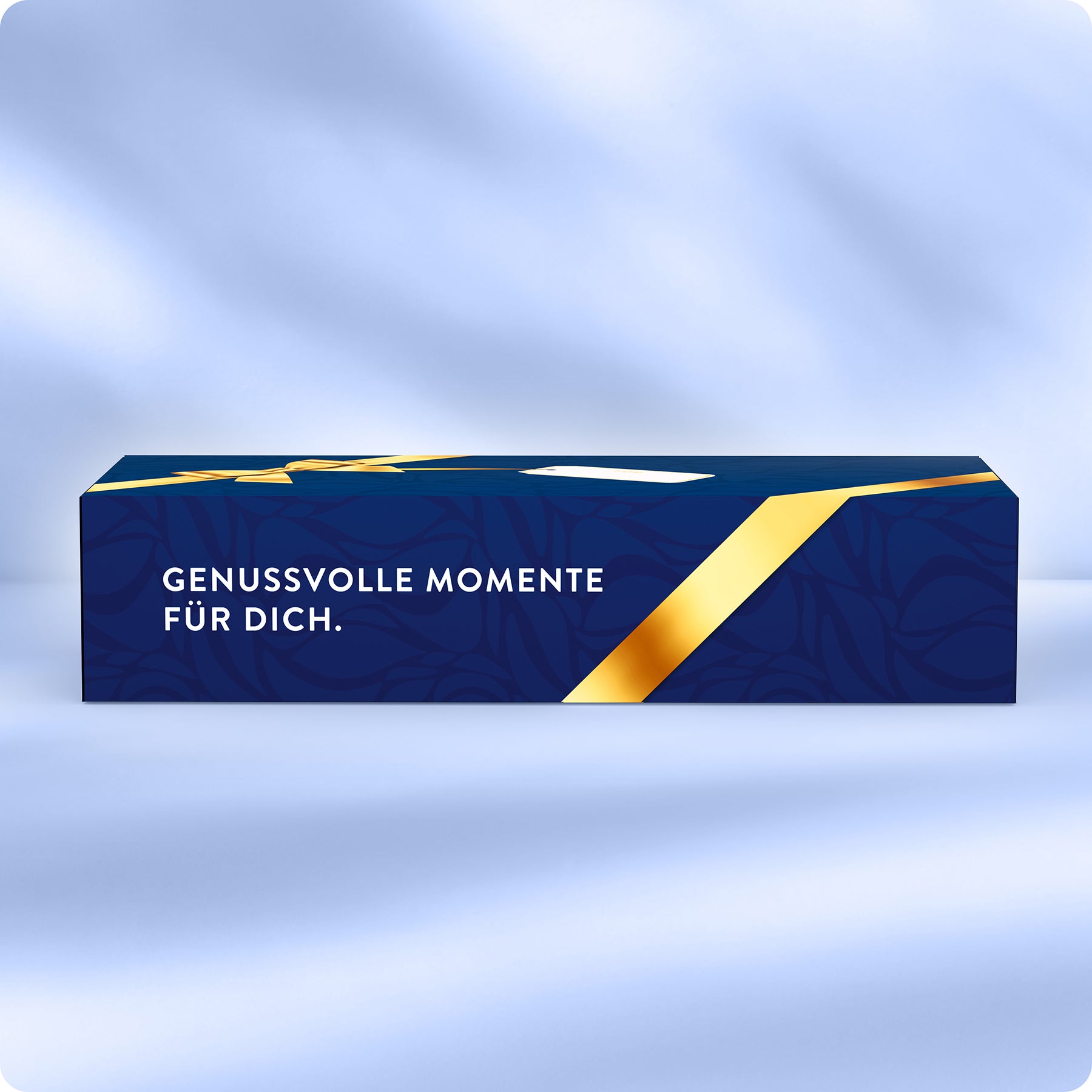 Eine rechteckige blaue Geschenkschachtel mit einem goldenen diagonalen Bandmuster und dem deutschen Text GENUSSVOLLE MOMENTE FÜR DICH. auf der Seite, vor einem sanften blauen Hintergrund.
