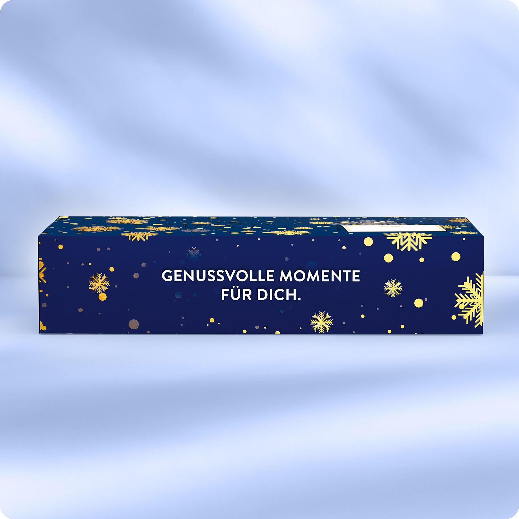 Eine rechteckige blaue Geschenkbox mit goldenen Schneeflocken und Punkten, die den deutschen Text GENUSSVOLLE MOMENTE FÜR DICH tragen. Die Schachtel ist vor einem weichen, hellblauen Hintergrund platziert.