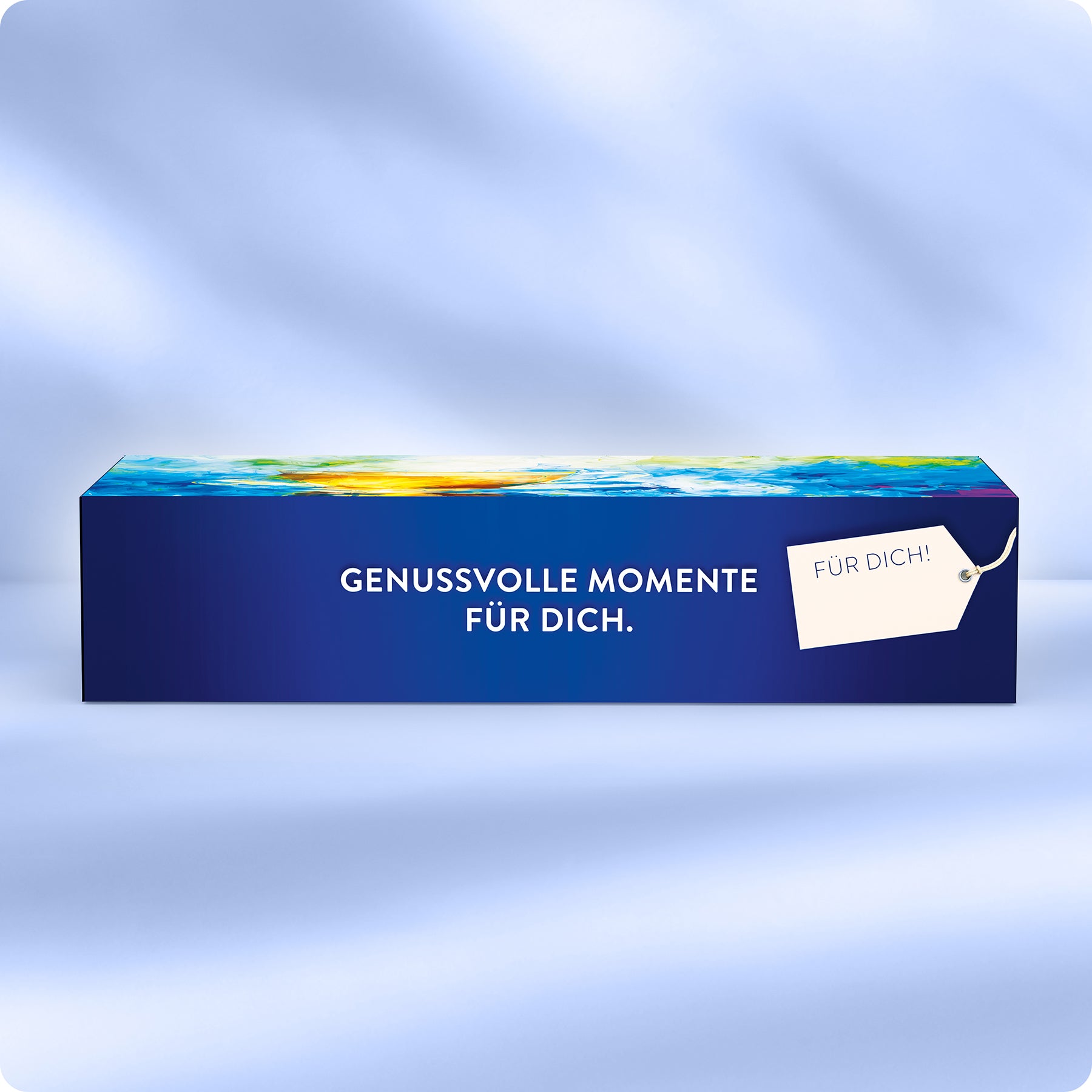 Eine rechteckige blaue Geschenkschachtel mit einem bunten oberen Rand, beschriftet mit GENUSSvolle MOMENTE FÜR DICH. Auf der rechten Seite befindet sich ein weißes Schild mit der Aufschrift FÜR DICH! Der Hintergrund ist weich und hellblau.