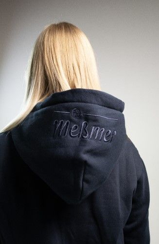 Eine Person mit glattem blondem Haar, von hinten gesehen, trägt einen gemütlichen Meßmer DE Hoodie mit dem aufgestickten Markennamen auf der Kapuze, vor einem schlichten, schwach beleuchteten Hintergrund.